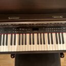 Digitální piano v tmavě hnědém dřevěném provedení. Na horní liště je nápis YAMAHA a pod ním ovládací panel s displejem a řadou tlačítek. Klaviatura m…
