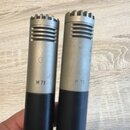 Dvě stříbrné kovové kondenzátorové kapsle mikrofonů s černým spodním tělem. Na obou je vyražen nápis M 73. Povrch kovové části vykazuje drobné škrába…