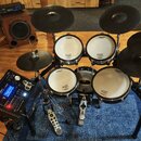 Elektronická bicí souprava značky Roland s bílými membránami. Soustava obsahuje čtyři padové bubny, tři činely a ovládací modul s nápisem Roland. Vše…