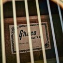 Fotografie zachycuje vnitřní štítek akustické kytary viditelný skrz otvor. Na bílém štítku s ozdobným rámem je uveden nápis Greco GUITARS, MODEL NO.…