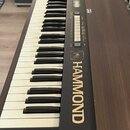 Elektronický klavír v hnědém dřevěném provedení s černým ovládacím panelem. Na boku je viditelný zlatý nápis HAMMOND a v horní části označení modelu…