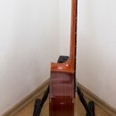 Fotografie zachycuje akustickou kytaru z profilu, umístěnou na černém stojanu. Nástroj má hnědý lakovaný povrch s viditnou strukturou dřeva. Na hlavě…