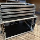 Dvě hliníkové transportní bedny s černým povrchem, naskládané na sebe. Bedny mají kovové rohové zpevnění a hliníkové profily s viditelnými šrouby. Vn…