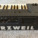 Černý klavír s bílými a černými klávesami. Na zadní straně je nápis KURZWEIL a čtyři konektory s popisky MIDI IN, MIDI THRU, MIDI OUT a SCSI. Povrch…