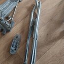 Na dřevěné podlaze leží tři kovové části pedálů v stříbrné barvě. Jsou vidět dva šlapací mechanismy s žebrovaným povrchem a jedna samostatná kovová p…