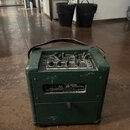 Zelený kytarový kombo zesilovač s černou rukojetí. Na ovládacím panelu je logo VOX a nápisy VOL VOLUME, TONE, GAIN a REVERB. Na zadní straně je štíte…