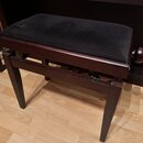 Tmavně hnědá klavírní stolička s černým čalouněným sedákem. Pod sedákem je vidět kovový mechanismus pro regulaci výšky a na boku dřevěný úchyt. Nástr…