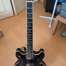 Černá polodublová elektrická kytara s bílým bindingem po obvodu těla a hradla. Na hlavě je viditelné logo HB. Nástroj má dva humbuckery, dva ovládací…