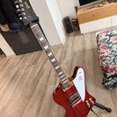 Červená elektrická kytara s bílým pickguardem a hnedým hmatníkem. Na hlavě je zlatý nápis Gibson, na těle se nachází bílý štítek s červeným logem a n…