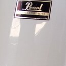 Detail bílého, lesklého povrchu bubnu. Na něm je umístěna obdélníková nálepka s černým pozadím a bílým nápisem Pearl a PEARL EXPORT SERIES. Povrch je…