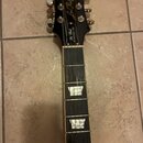 Černá kytara s hnedým hmatníkem a trapezoidovými vložkami. Na hlavě je bílý nápis Epiphone a zlatý nápis Les Paul. Pod ním je uveden model Les Paul S…