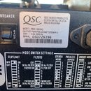 Detailní záběr na zadní panel zesilovače s bílou nálepkou, na které je logo QSC a nápisy: QSC AUDIO PRODUCTS COSTA MESA, CA USA MADE IN CHINA. Je zde…