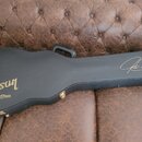 Černý tvrdý kufr na kytaru s zlatými detaily a úchytem. Na povrchu je zlatý nápis Gibson Custom a v pravé části stylizovaný podpis. Kufr vykazuje mír…