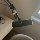 Na fotografii je černý mikrofon s nasazenou šedou větrolnicí, který je upevněn na nastavitelné rameni. Na těle mikrofonu je viditelný symbol CE a neč…