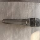 Tmavě šedý dynamický mikrofon s kovovou síťkovou hlavicí. Na těle je vidět bílý nápis SHURE a v dolní části označení KSM9. Povrch mikrofonu je matný…
