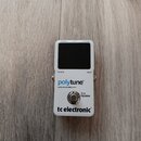 Bílý kytarový ladič s černým displejem. Na přední straně jsou nápisy „display“, „pitch“, „tuning“, „output“ a „input“. Uprostřed je modrý a černý náp…
