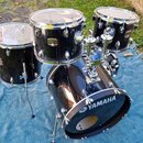 Černá bubnová sada s kovovým hardwarem, umístěná na modré přikrévce. Na basovém bubnu je bílý nápis YAMAHA s doprovodným logem. Na tomu, který je umí…