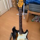 Elektrická kytara s tělem v provedení sunburst a bílým pickguardem. Na hlavě hředu je viditelný nápis „by Tussock“. Kytara má hnědý hmatník a je umís…