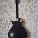 Zadní strana černé elektrické kytary s tvarem singlecut. Na zadní straně těla je nalepený čtvercový štítek s logem a nápisem DUNLOP STRINGS. Kytara m…