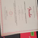 Fotografie zobrazuje certifikát pravosti a červený papírový obal s logem Fender. Na certifikátu je uveden nápis CERTIFICATE of AUTHENTICITY, značka F…