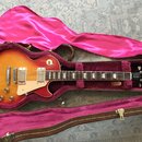 Elektrická kytara s tónem sunburst v hnědo-žlutých odstínech, umístěná v otevřeném hnědém kufru s fialovou výplní. Na hlavě kytary je bílý nápis Gibs…