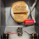 Interiér šedého kovového šasi s elektronickými komponenty. Uprostřed je velký stříbrný válečný kondenzátor přelepený hnědou lepicí páskou, vedle něj…