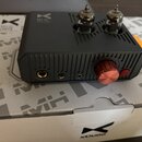 Šedý stolní zesilovač s hliníkovým žebrovaným šasi a dvěma viditelnými elektronkami. Na horní části je nápis MH-02 a logo xDuoo, na přední straně se…