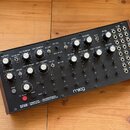 Černý semi-modulární analogový perkusní syntetizér v dřevěném šasi. Na předním panelu je nápis DFAM DRUMMER FROM ANOTHER MOTHER SEMI-MODULAR ANALOG P…