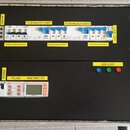 Elektrický rozvaděč v kovovém kufru s černým dnem. Horní část obsahuje jističe a štítky s nápisy PHASE, Fi SCHALTER-OUT1, Fi SCHALTER-OUT2, LED LICHT…