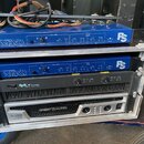Krytý rack s audio zařízeními. Obsahuje dva modré zesilovače s nápisy NEXO, LSub 500 a PS10. Pod nimi je černý zesilovač s nápisem Zeck PT 7 POWER TR…