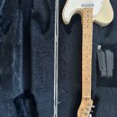 Kytara s krémovým tělem a javorovým hnedem, položená v černém plišovém kufru. Na hlavě je viditelné logo Fender a nápis TELECASTER. Vedle krku kytary…