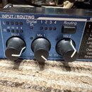 Detailní záběr na modrý panel efektového procesoru s nápisy INPUT / ROUTING a REVERB. Jsou zde tři černé otočné ovladače s bílými značkami popsané ja…