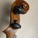 Detail hlavy smyčcového nástroje z hnědého dřeva s ozdobnou spirálou. Jsou vidět dva černé ladicí kolíky s oválnými hlavami. Povrch dřeva vykazuje dr…