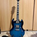 Modrá elektrická kytara s tvarem SG, umístěná na stojanu. Na hlavě hředu je nápis Epiphone. Kytara má dva humbuckery, tři ovládací knoflíky a stрійko…