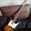 Elektrická kytara s tělem v barvě sunburst a bílým pickguardem. Na hlavě hředu je viditelné logo Squier. Kytara leží na černé softcase kytarové tašce…