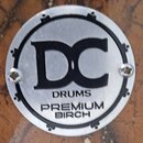 Kruhová kovová plákovka s černými nápisy a grafickými prvky. Uprostřed je velké logo DC, pod kterým jsou slova DRUMS a PREMIUM BIRCH. Plákovka je při…