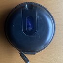 Černý kulatý nabíjecí box v textilním pouzdře. Na horní části je vidět svítící modrá kontrolka a symbol baterie. Na stahovadle zipu je nápis SHURE. P…