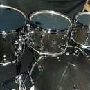 Sada černých bubnů s chromovaným kováním. Na jednom z tomů je viditelné logo Sonor. V pozadí jsou cardboardové krabice s nápisem SONOR Made in German…