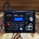 Černý elektronický modul k bubnům značky Roland s nápisem DRUM SOUND MODULE TD-30 SuperNATURAL. Zařízení má digitální displej s modrým podsvícením a…