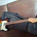 Elektrická kytara s tělem v barvě sunburst a bílým pickguardem, položená na černém softcasu. Na hlavě hředu je viditelné logo „Squier“. Hmatník je vy…
