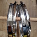 Dvě chromované kovové obruče na buben, které jsou opřené o dřevěnou paličku. Na obou je viditelný bílý štítek s nápisy MAPEX 2.3mm POWER HOOPS. Povrc…