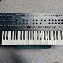 Široký analogový synthesizér s klaviaturaí a dřevěným bočním rámem. Horní panel je šedý s mnoha ovládacími knoflíky a přepínači, rozdělenými do sekcí…