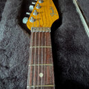 Hlava a hриф elektrické kytary s tmavým hnedým hmatníkem. Na žluté hlavě je černý nápis Fender® STRATOCASTER MADE IN USA a sériové číslo E 919409. Ná…