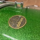 Detailní záběr na zelený buben s glitrovým povrchem. Na korpusu je připevněna kovová oválná plákovka s nápisy „Dios SERIES DRUMS ddrum Hand-Crafted S…