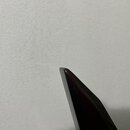 Detailní záběr na špičku tmavě červeného, lakovaného předmětu s hladkým povrchem. Předmět je umístěn před bílou, strukturovanou stěnou. Žádné značky…