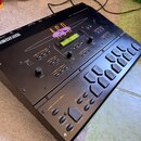Černý sampler Ensoniq ASR-10 s LCD displejem a gumovými pady. Na horní části je bílý nápis ENSONIQ v rámečku a velký oranžový nápis ASR-10. Zařízení…