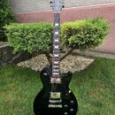 Černá elektrická kytara s tvarem single-cutaway a hnědým hmatníkem. Na hlavě je viditelné logo Gibson a nápis Model. Tělo kytary má dva humbuckery, č…