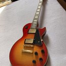 Elektrická kytara s trupem v červeno-žlutém sunburst provedení a zlatým hardwarem. Na hlavě hředu je viditelné logo Gibson. Kytara má dva humbuckery,…