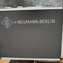 Černý přepravní kufr s hliníkovým rámem a kovovými zámky. Na čelní straně je bílý nápis NEUMANN.BERLIN a vedle něj logo značky Neumann v kosočtverci.…