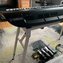 Černý digitální klavír s bílými a černými klávesami, umístěný na bílém kovovém stojanu. Na levé straně korpusu je bílé logo YAMAHA. V zadní části jso…